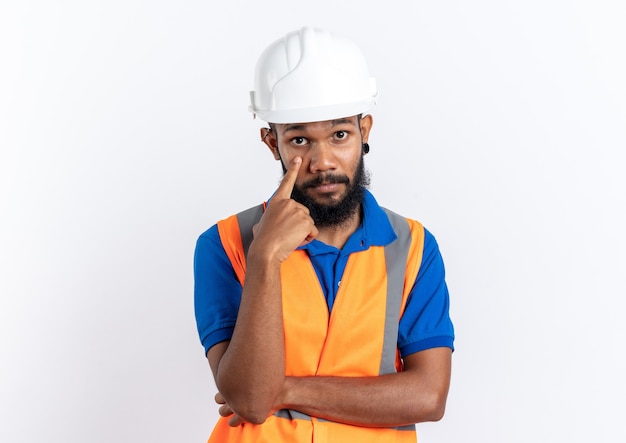 Confident young afro-american builder man in uniform with safety helmet pulling down eyelid with finger isolated on white wall with copy space