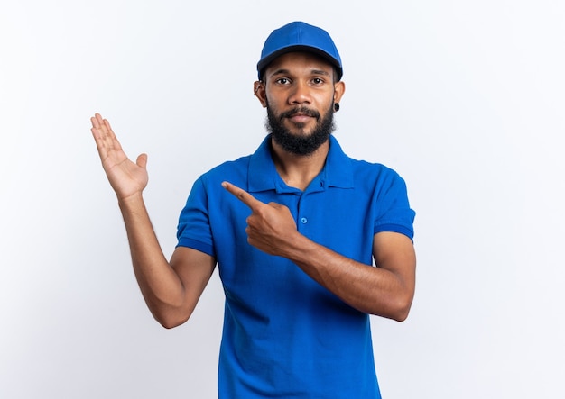 Confident young afro-american delivery man pointing at his hand isolated on white wall with copy space