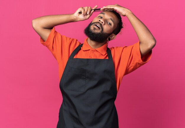 Confident young afro-american male barber wearing uniform  combing his hair isolated on pink wall