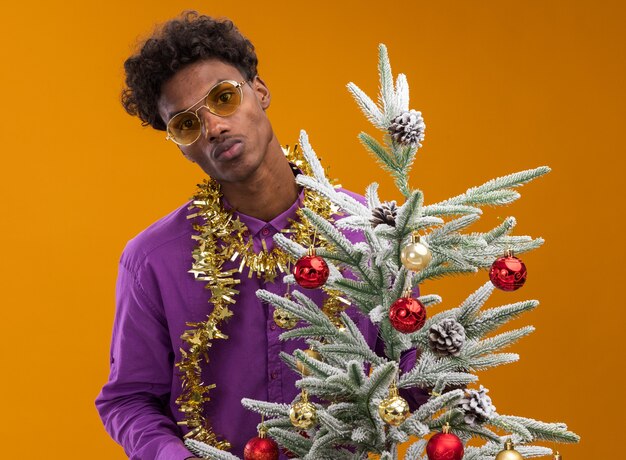 Confident young afro-american man wearing glasses with tinsel garland around neck standing behind decorated christmas tree  with pursed lips isolated on orange wall