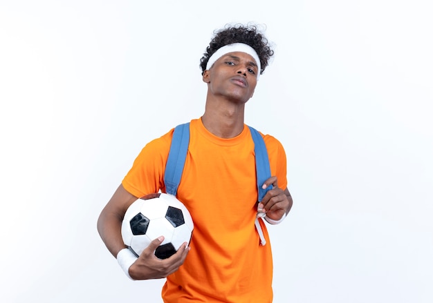 Confident young afro-american sporty man wearing headband and wristband and back bag holding ball isolated on white background