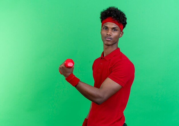 Confident young afro-american sporty man wearing headband and wristband exercising with dumbbell isolated on green background