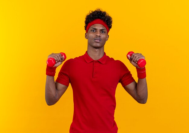 Уверенный молодой афро-американский спортивный мужчина с повязкой на голову и браслетом, тренирующимся с гантелями, изолированными на желтой стене