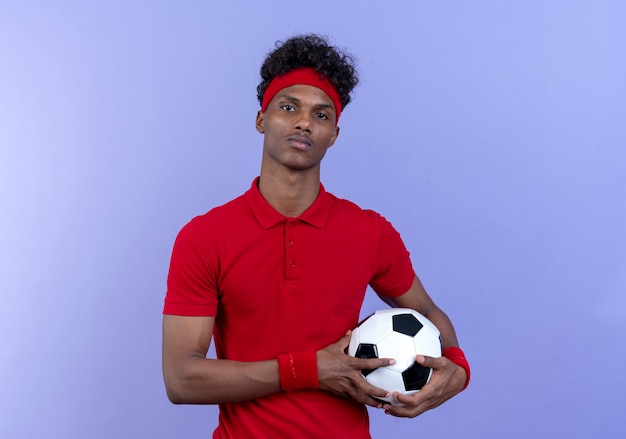 Free photo confident young afro-american sporty man wearing headband and wristband holding ball isolated on blue wall