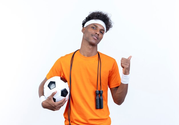 Confident young afro-american sporty man wearing headband and wristband holding ball with jump rope on shoulder and points at side isolated on white background with copy space