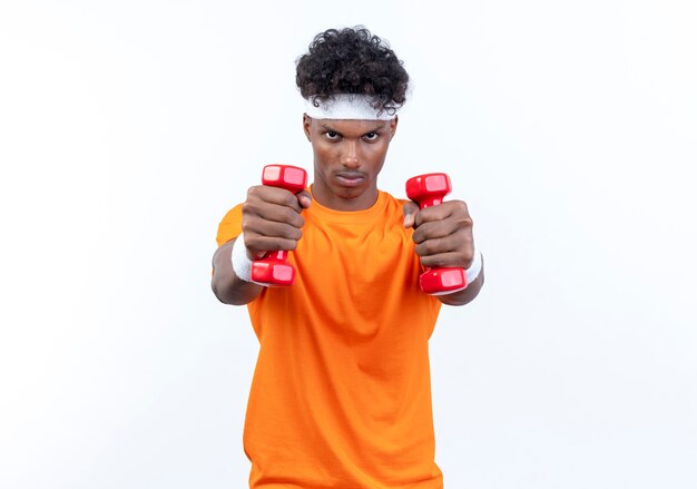 Confident young afro-american sporty man wearing headband and wristband holding out dumbbells