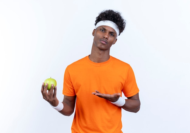 Confident young afro-american sporty man wearing headband and wristband holding and points with hand at apple 
