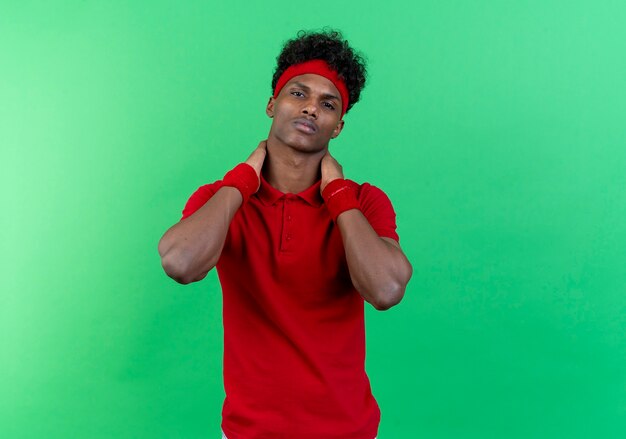 Confident young afro-american sporty man wearing headband and wristband keeping hand behind neck isolated on green background