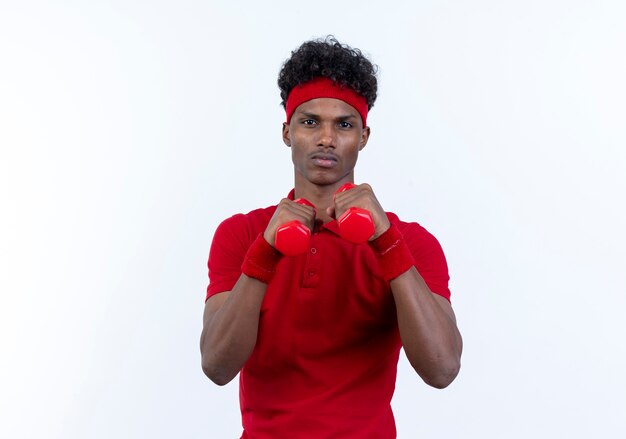 Confident young afro-american sporty man wearing headband and wristband standing in fighting pose and holding dumbbells isolated on white background