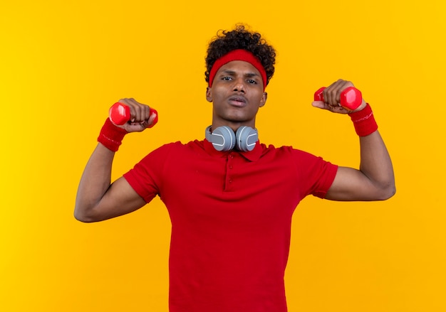 Confident young afro-american sporty man wearing headband and wristband with headphones on neck raising dumbbell isolated on yellow background