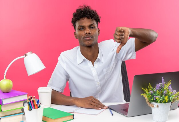 Confident young afro-american student sitting at desk with school tools thumbing down isolated on pink wall