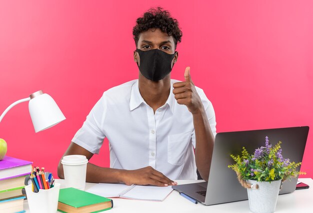 Confident young afro-american student wearing face mask sitting at desk with school tools thumbing up 