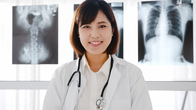 청진 기 카메라를보고 하 고 건강 병원에서 환자와 화상 회의 통화하는 동안 웃 고 흰색 의료 제복을 입은 자신감이 젊은 아시아 여성 의사.