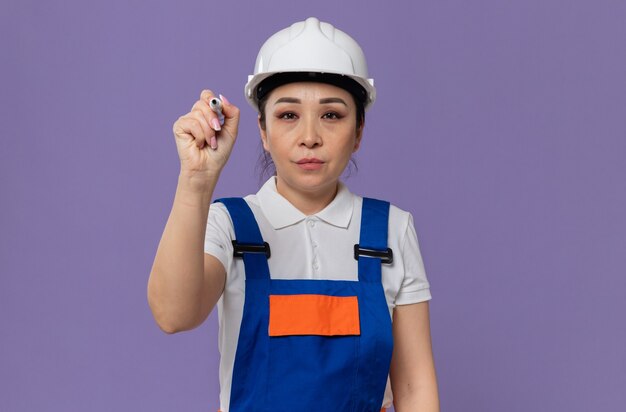 マーカーを保持している白い安全ヘルメットを持つ自信を持って若いアジアのビルダーの女の子