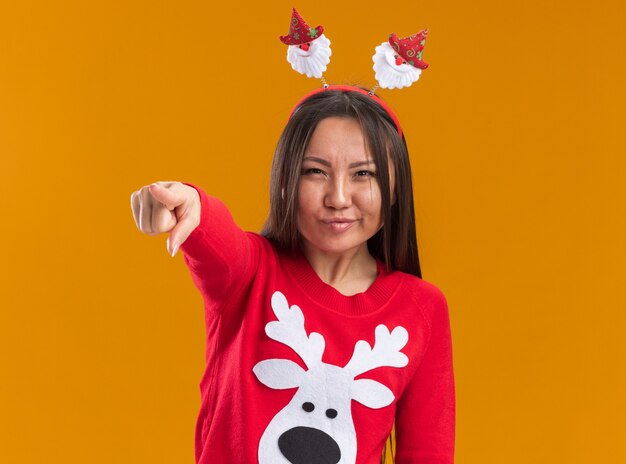 Уверенная молодая азиатская девушка в рождественском обруче для волос со свитером, показывающим вам жест, изолированный на оранжевом фоне
