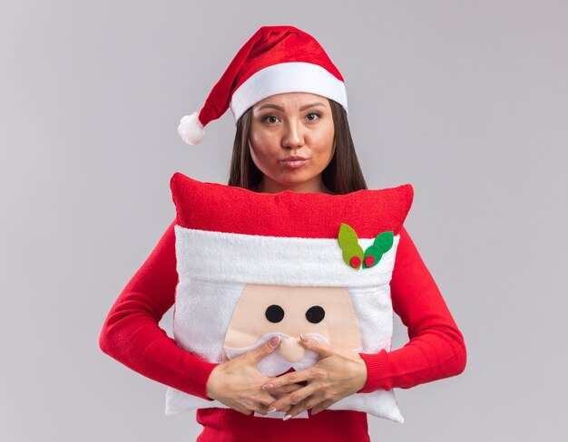 白い背景で隔離のクリスマス枕を保持しているセーターとクリスマス帽子をかぶって自信を持って若いアジアの女の子