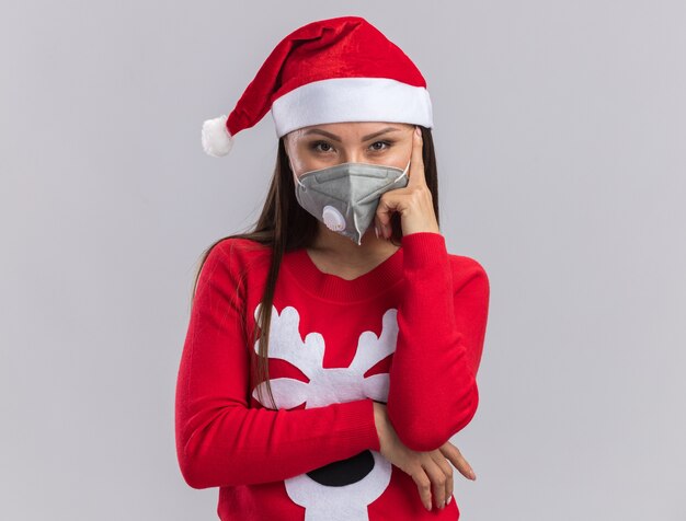 白い背景で隔離の頬に指を置くセーターと医療マスクとクリスマス帽子を身に着けている自信を持って若いアジアの女の子