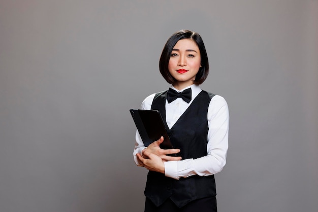 デジタルタブレットの肖像画を持って立っているユニフォームと蝶ネクタイを着た自信のある若いアジア人女性ウェイトレス ⁇ 携帯ガジェットを手に持ってカメラを見ている深刻なレセプショニスト