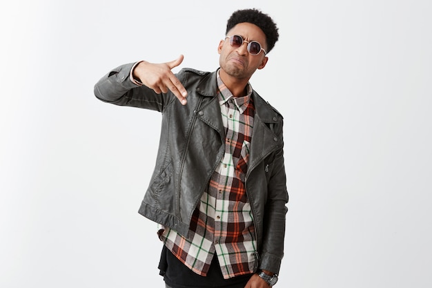 Confident young attractive black-skinned african male with curly hair in trendy leather jacket and sunglasses making gun gesture with hand with mean face expression.