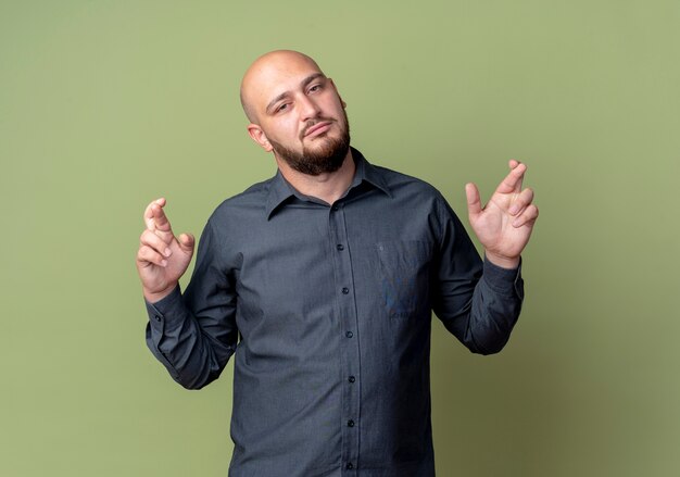 Confident young bald call center man doing crossed fingers gesture isolated on olive green 