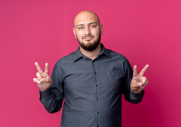 Confident young bald call center man doing peace signs isolated on crimson 