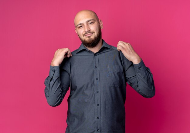 Confident young bald call center man grabbing his shirt isolated on crimson  with copy space