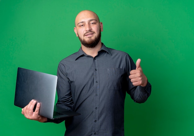 Confident young bald call center man holding laptop and showing thumb up isolated on green  with copy space