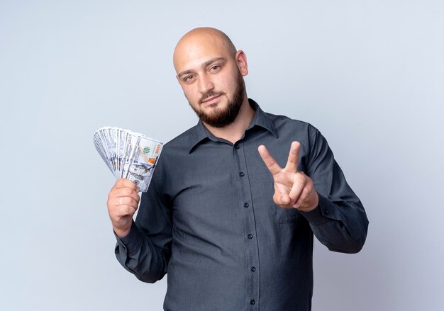 Confident young bald call center man holding money and doing peace sign isolated on white 