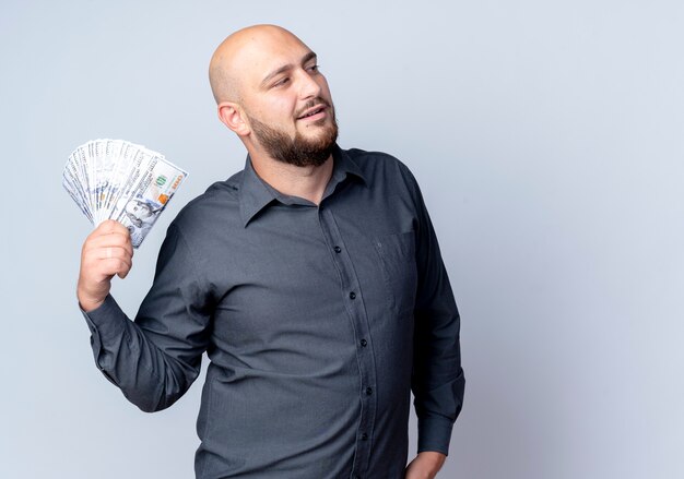 Confident young bald call center man holding money looking at side isolated on white  with copy space