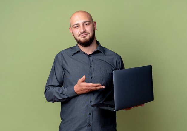 Confident young bald call center man holding and pointing with hand at laptop isolated on olive green  with copy space