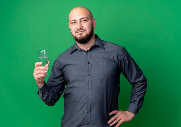 Confident young bald call center man keeping hand on waist and holding glass of water isolated on green 