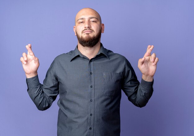 Confident young bald call center man looking doing crossed fingers gesture isolated on purple 
