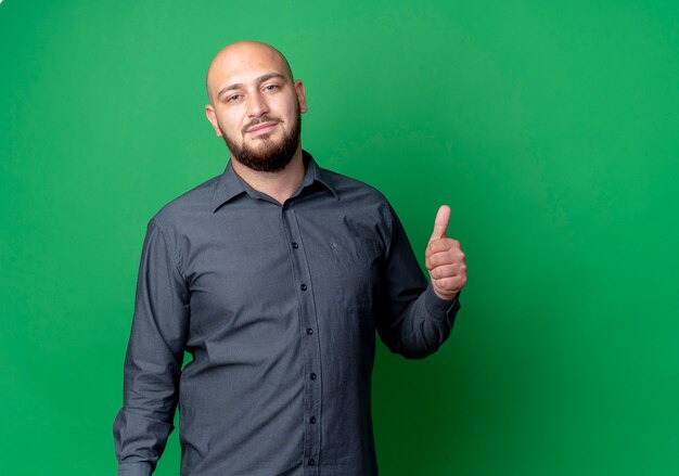 Confident young bald call center man looking and showing thumb up isolated on green  with copy space