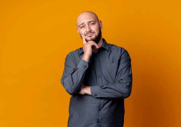 Confident young bald call center man putting hand on chin looking isolated on orange  with copy space