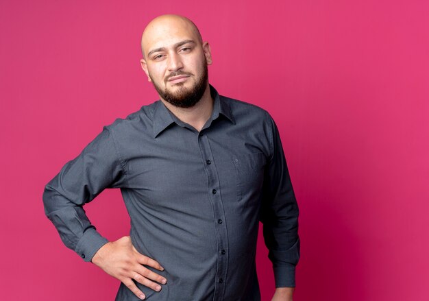 Confident young bald call center man putting hand on waist isolated on crimson  with copy space