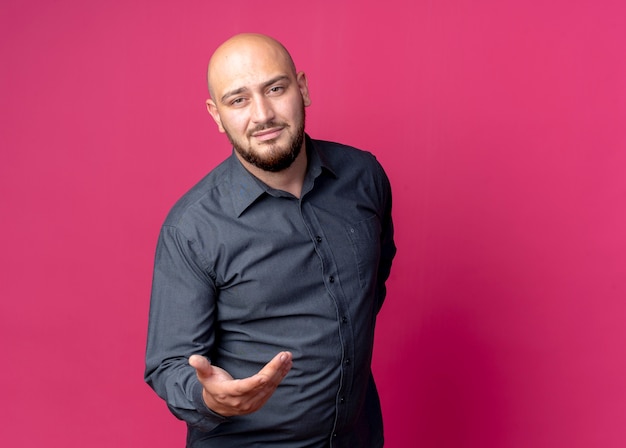 Confident young bald call center man showing empty hand and keeping another hand behind back isolated on crimson  with copy space