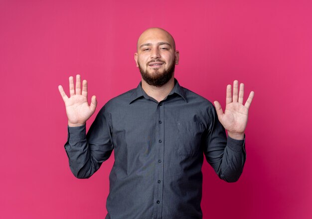 Confident young bald call center man showing empty hands isolated on crimson 