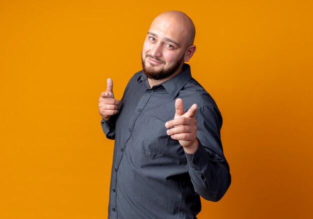 Confident young bald call center man standing in profile view doing you gesture isolated on orange  with copy space