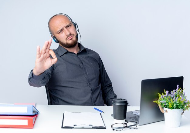 Confident young bald call center man wearing headset sitting at desk with work tools doing ok sign isolated on white 