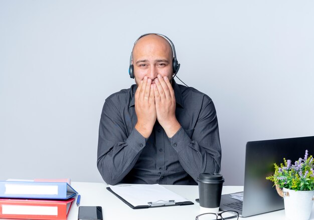 Confident young bald call center man wearing headset sitting at desk with work tools putting hands on mouth isolated on white 