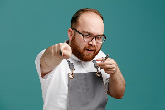 Confident young barber wearing uniform and glasses holding scissors and comb looking at camera showing you gesture with comb and scissors isolated on blue background