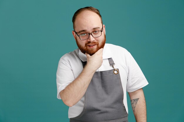 Confident young barber wearing uniform and glasses looking at camera while keeping hand on chin isolated on blue background