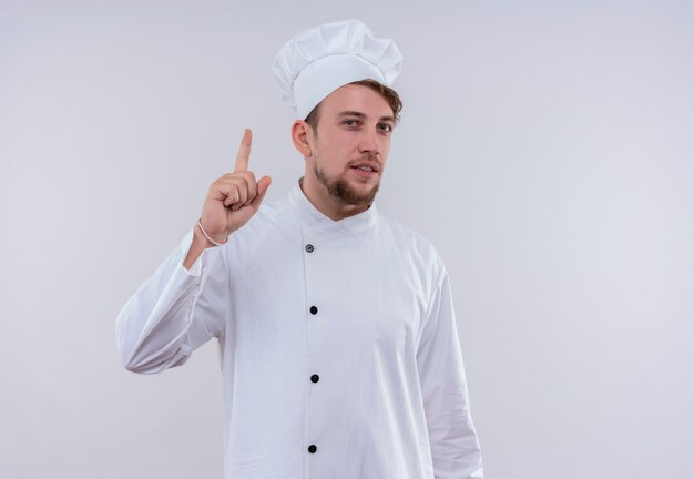 A confident young bearded chef man wearing white cooker uniform and hat pointing up with index finger while looking on a white wall