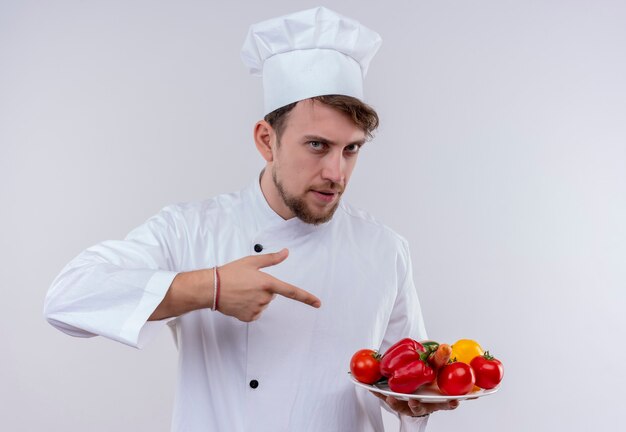 白い炊飯器の制服と白い壁に新鮮な野菜と白いプレートを指す帽子を身に着けている自信を持って若いひげを生やしたシェフの男