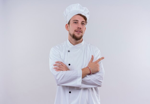 A confident young bearded chef man wearing white cooker uniform and hat pointing with index finger and holding hands folded while looking on a white wall