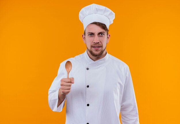 A confident young bearded chef man in white uniform holding wooden spoon while looking on an orange wall