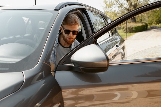 Уверенный молодой бородатый мужчина в модных солнцезащитных очках за рулем автомобиля.