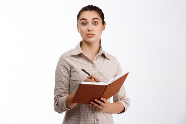 Confident young beautiful brunette businesswoman writing in notebook over white wall