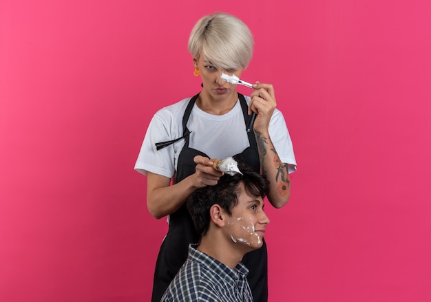 Foto gratuita fiducioso giovane e bella barbiere femminile in uniforme che tiene strumenti da barbiere e si fa la barba per un ragazzo isolato sul muro rosa