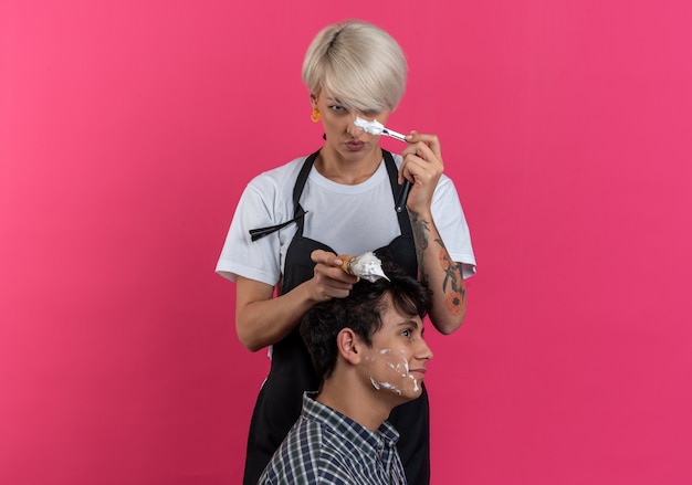 Confident young beautiful female barber in uniform holding barber tools and doing beard shaving for guy isolated on pink wall